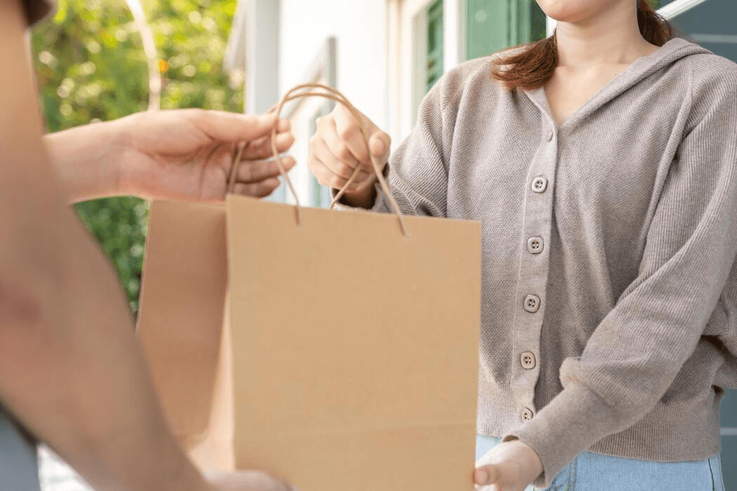 Kurir menyerahkan kantong belanja kraft ke tangan pelanggan di depan rumah